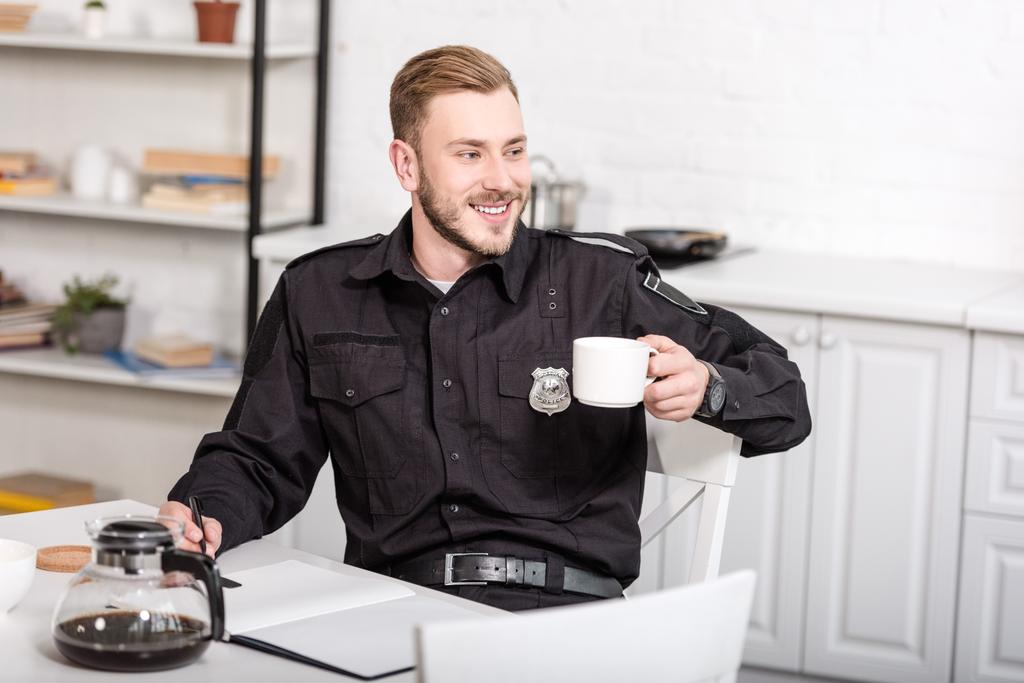 красивый офицер полиции сидит за кухонным столом, улыбается и пьет кофе
 - Фото, изображение
