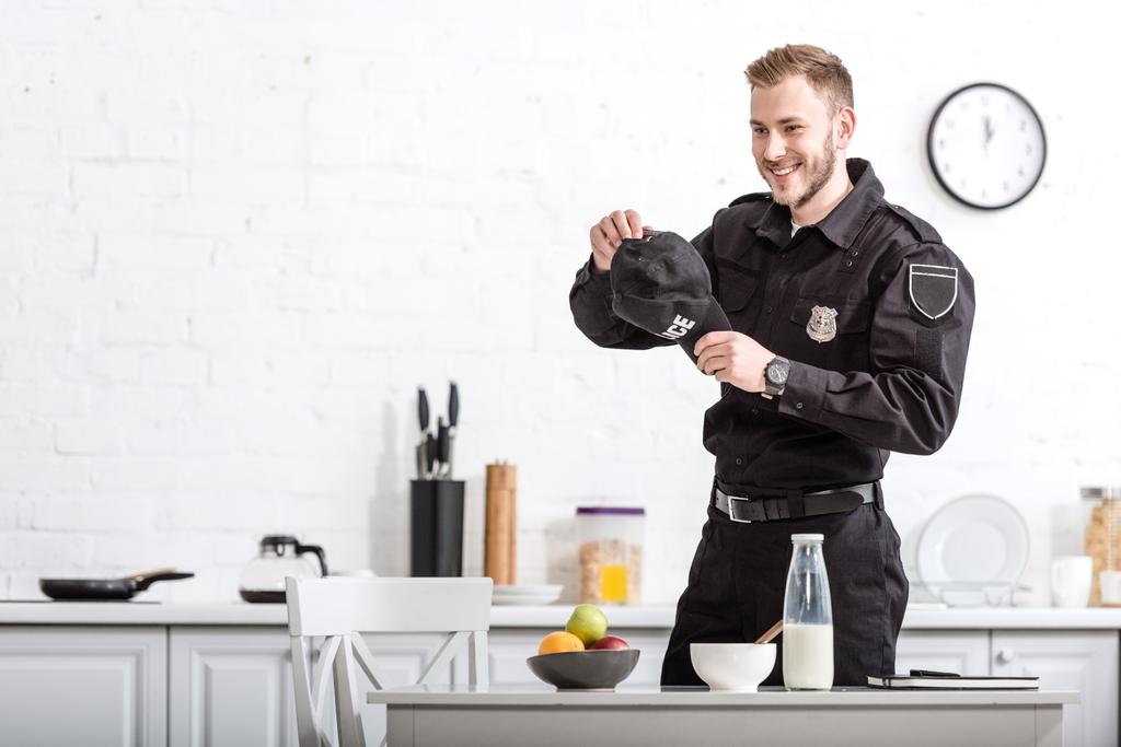 schöner Polizeibeamter mit Mütze in den Händen, der in die Küche lächelt - Foto, Bild