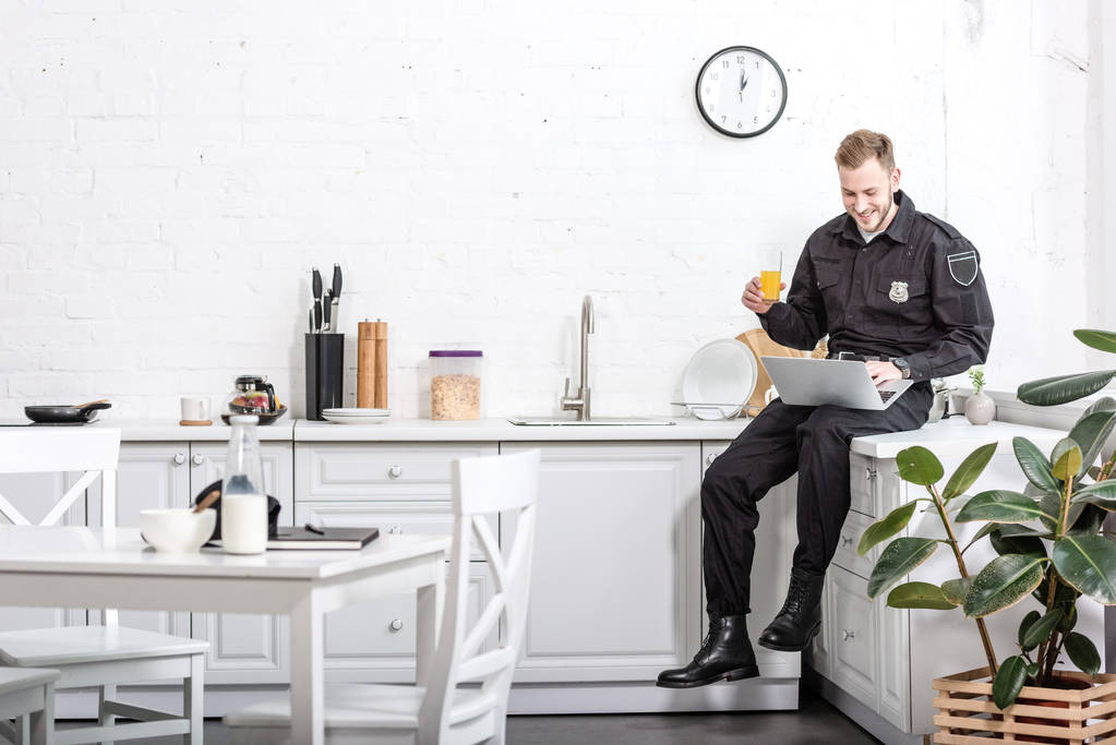 Young policeman sitting on table, drinking orange juice and using laptop at kitchen - Photo, Image
