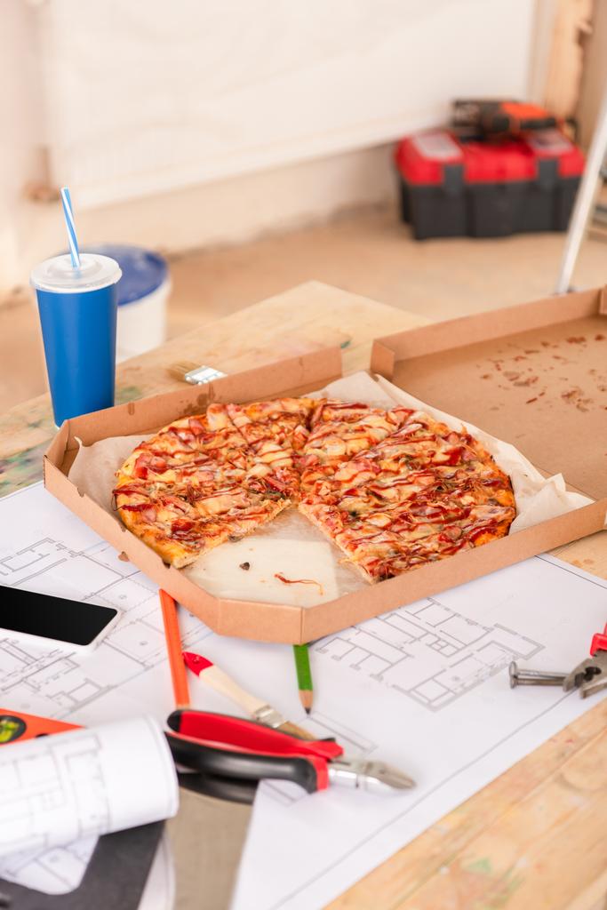 hoge hoekmening van pizza, frisdrank, blauwdruk, gereedschappen en smartphone met leeg scherm op tafel - Foto, afbeelding