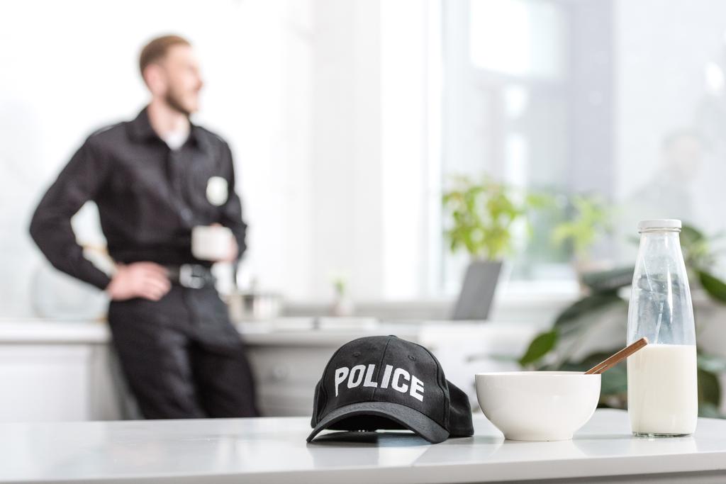 Polizeimütze, Schüssel und Milchflasche auf Küchentisch und Polizist im Hintergrund - Foto, Bild