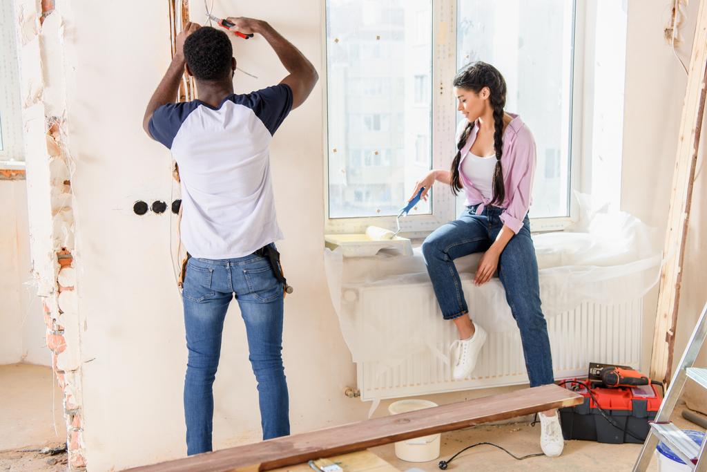 Задний вид человека, работающего с плоскогубцами, в то время как его подружка кладет ролик в роллер на подоконнике в новом доме
 - Фото, изображение