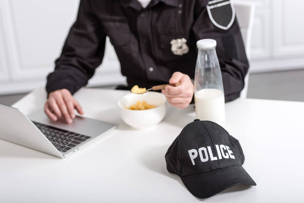 vista recortada del oficial de policía utilizando el ordenador portátil y comer copos de maíz en la mesa de la cocina
 - Foto, imagen