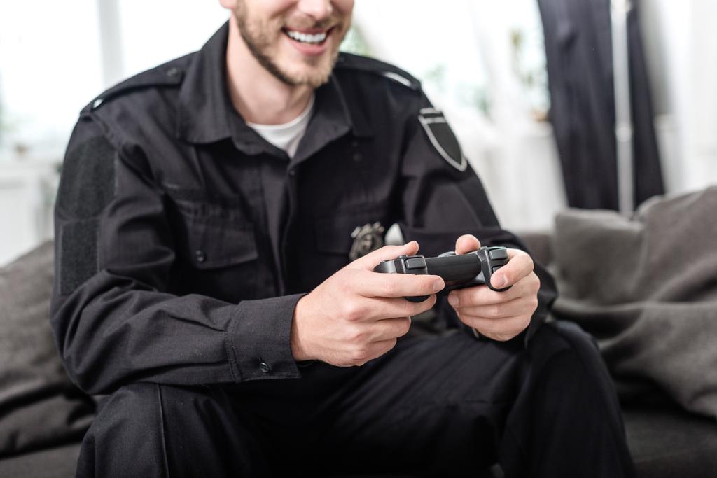 bijgesneden weergave van lachende politieman met gamepad zittend op de Bank - Foto, afbeelding