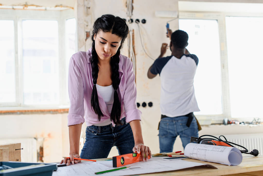задумчивая молодая женщина с уровнем духа глядя на план, в то время как ее парень работает позади во время ремонта дома
  - Фото, изображение