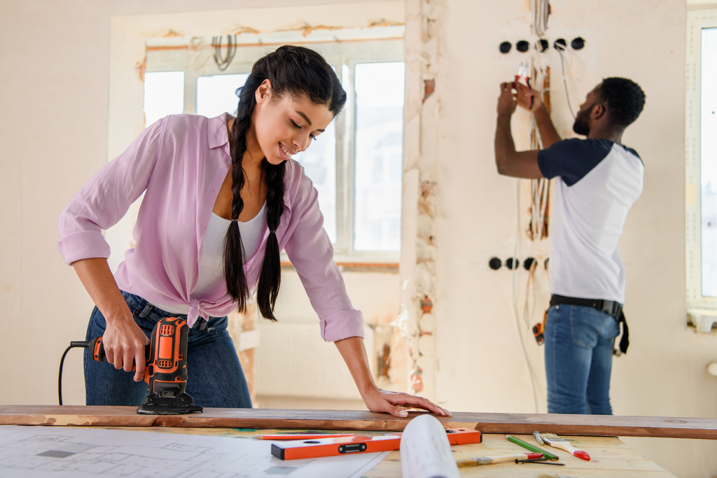 attractive african american woman working with jigsaw while her boyfriend standing behind during renovation at home - Photo, Image