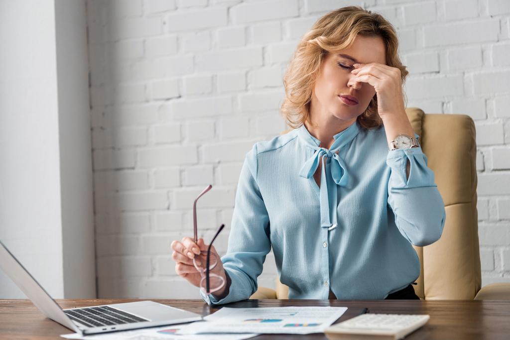 mujer de negocios cansada sosteniendo anteojos y frotando puente nasal en el lugar de trabajo
 - Foto, imagen
