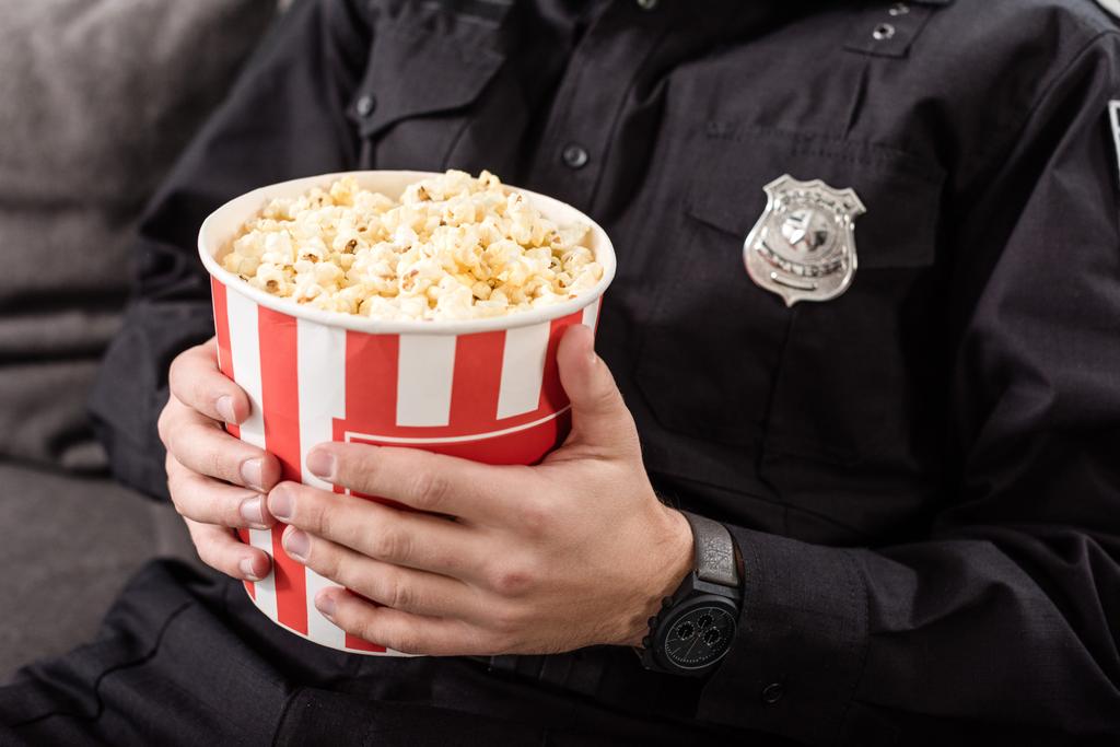 oříznutý pohled policista sedící na gauči s pruhovanou popcorn kbelík - Fotografie, Obrázek
