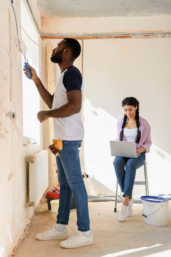 vista lateral de la pared de la pintura del hombre afroamericano mientras que su novia usando el ordenador portátil detrás durante la renovación del hogar
 - Foto, Imagen