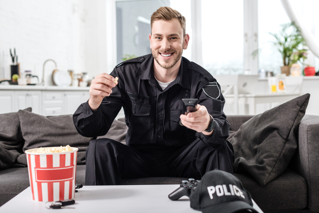 Офицер полиции сидит на диване, держит пульт дистанционного управления и ест попкорн
 - Фото, изображение