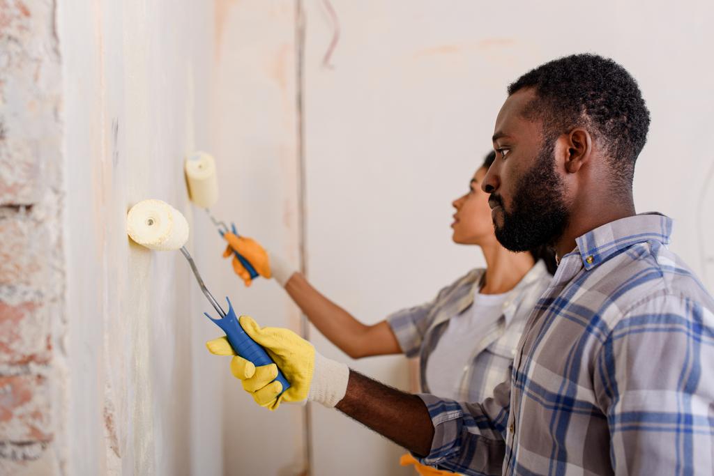 Seitenansicht eines afrikanisch-amerikanischen Paares beim Wandbemalen mit Farbwalzen im neuen Zuhause - Foto, Bild