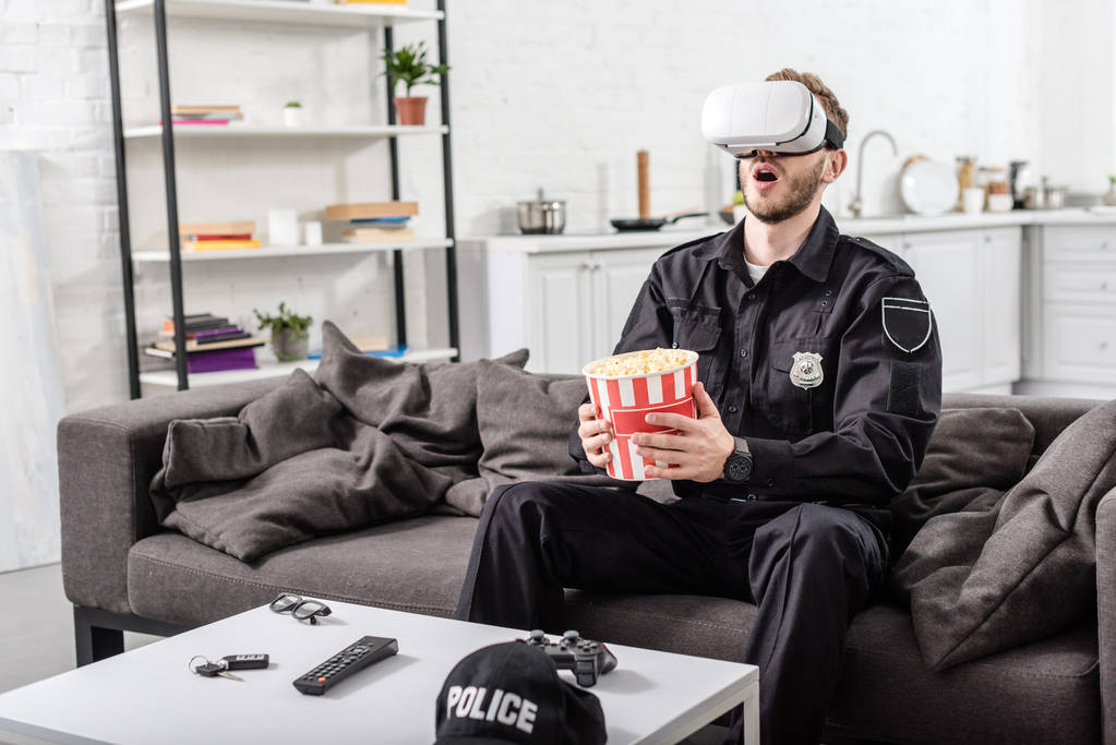 überraschter Polizeibeamter mit Virtual-Reality-Headset auf dem Kopf hält gestreiften Popcorn-Eimer und sitzt auf Couch  - Foto, Bild