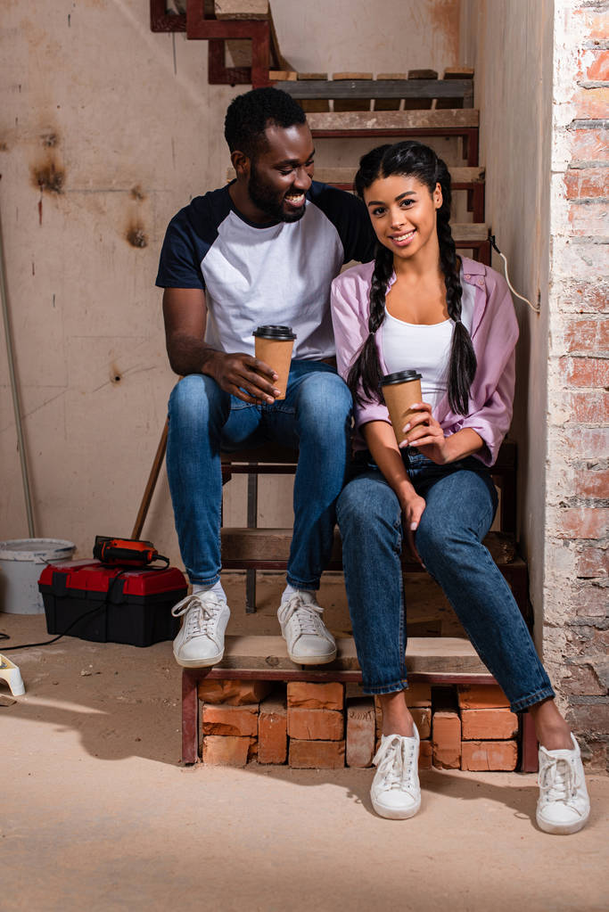 vrolijke Afrikaanse Amerikaanse echtpaar rusten met wegwerp kopjes koffie tijdens renovatie nieuwe thuis - Foto, afbeelding