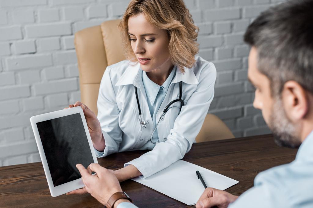 atractiva doctora mostrando tableta con pantalla en blanco al paciente en el consultorio
 - Foto, Imagen