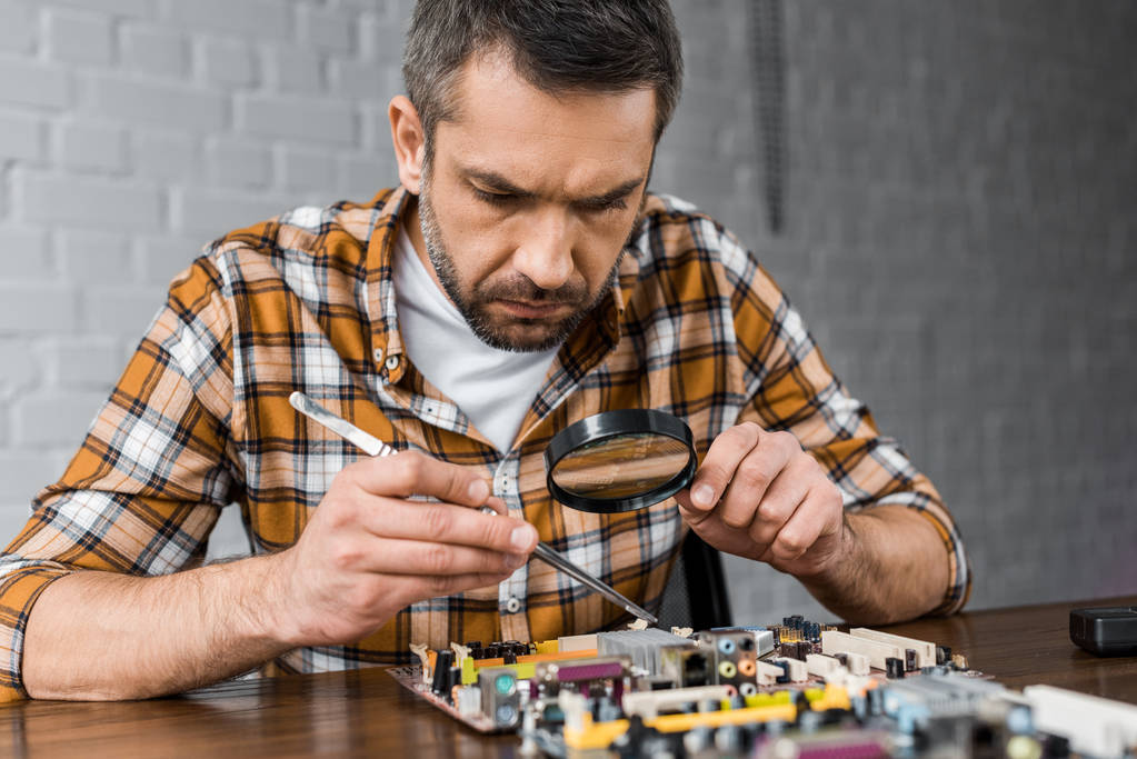 close-up portrait of concentrated electronics engineer with tweezers and magnifying glass repairing motherboard - Photo, Image