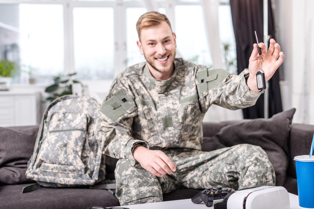 взволнованный солдат армии сидя на диване дома, глядя на камеру и держа ключ
 - Фото, изображение