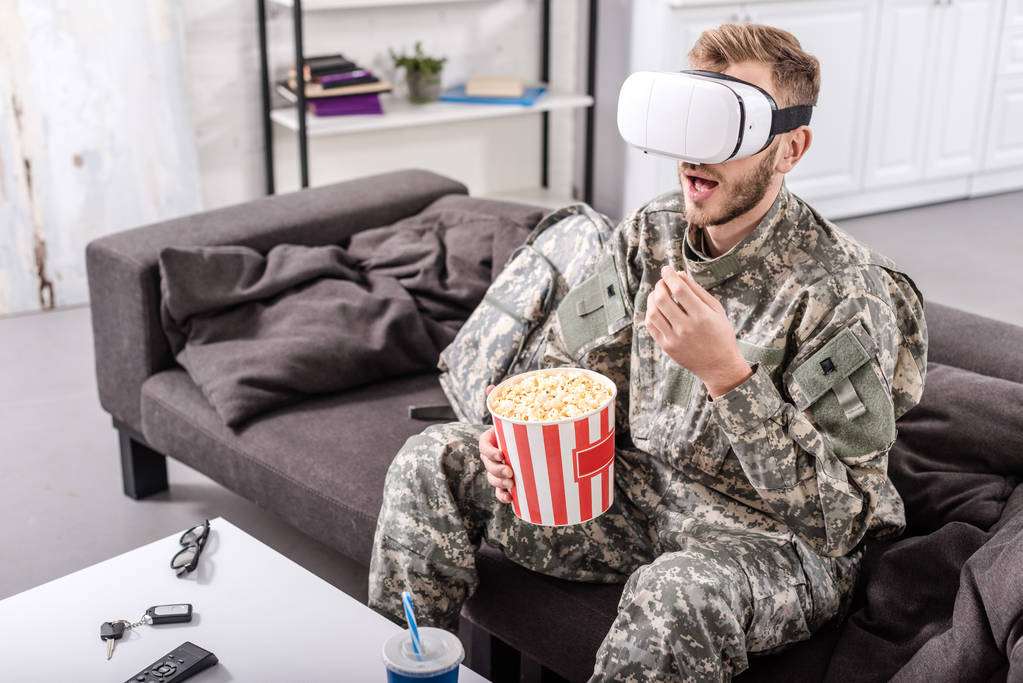 στρατιώτης φοράει σετ κεφαλής εικονικής πραγματικότητας, κάθεται στον καναπέ, παρακολουθώντας την ταινία και τρώγοντας ποπ κορν - Φωτογραφία, εικόνα