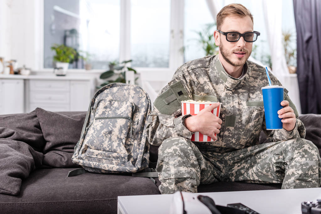 knappe soldaat in 3D-bril op bank kijken naar film met popcorn en soda water - Foto, afbeelding