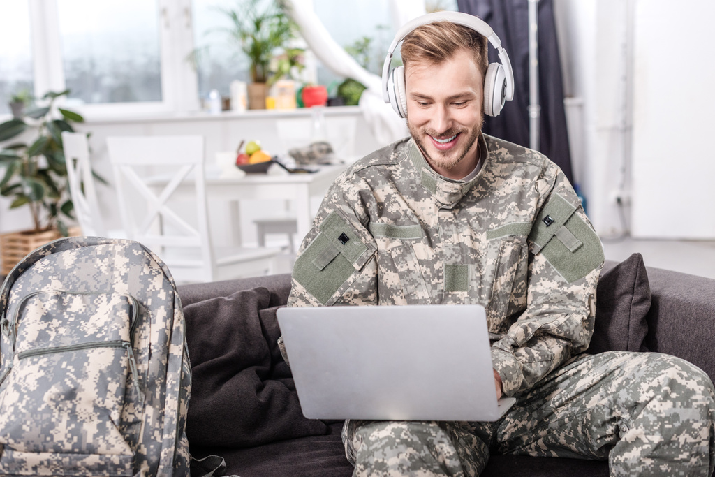 χαμογελώντας στρατού στρατιώτης φορώντας ακουστικά και χρησιμοποιώντας το laptop στον καναπέ - Φωτογραφία, εικόνα