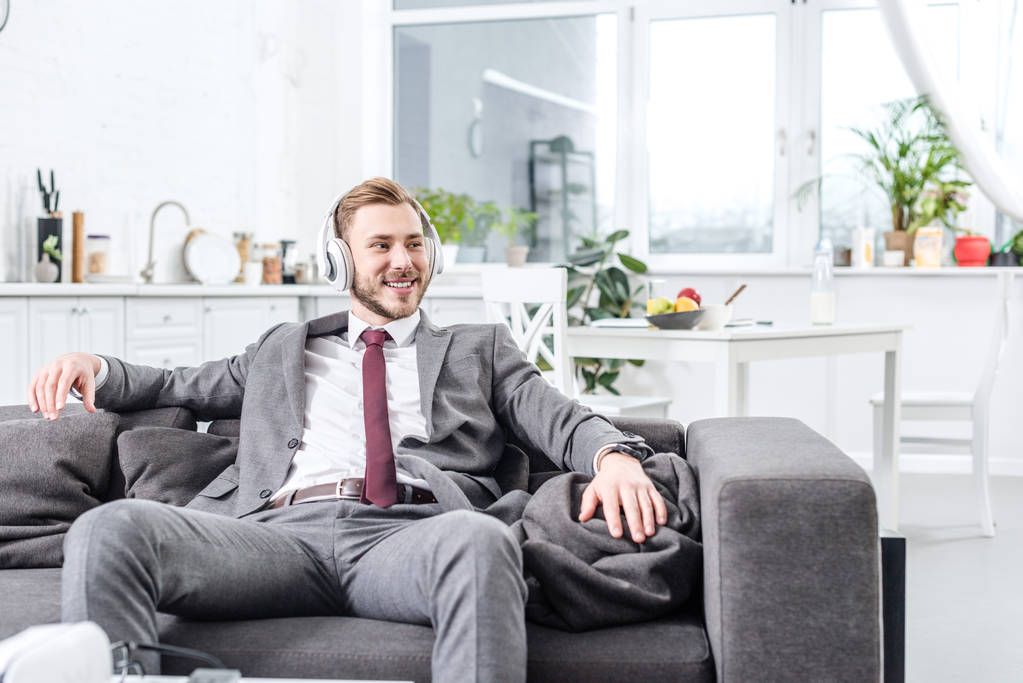 Улыбающийся бизнесмен в формальной одежде в наушниках и сидящий дома на диване
 - Фото, изображение