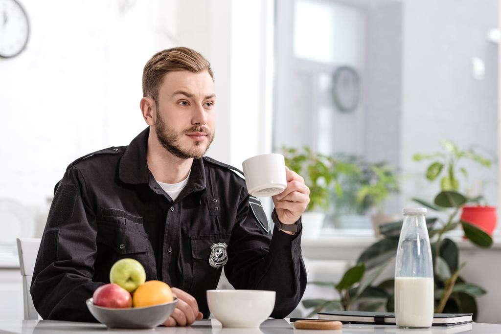policista v kuchyni sedí, drží šálek horké kávy a po snídani v kuchyni u stolu - Fotografie, Obrázek