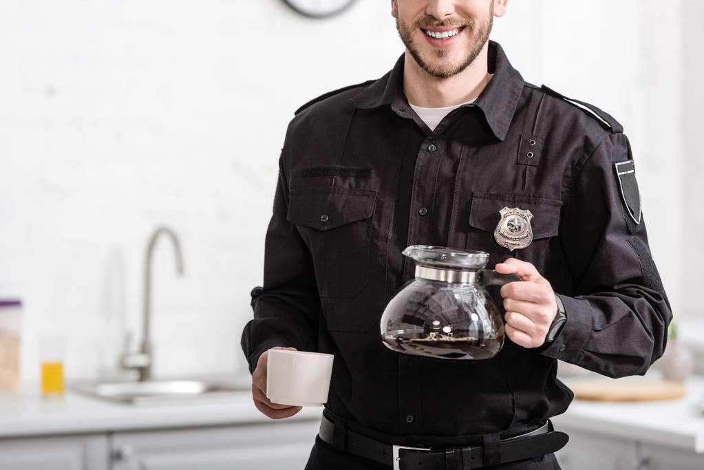 Μερική άποψη της χαμογελώντας αστυνομικός κρατώντας γυάλινο δοχείο του φιλτραρισμένου καφέ στην κουζίνα - Φωτογραφία, εικόνα