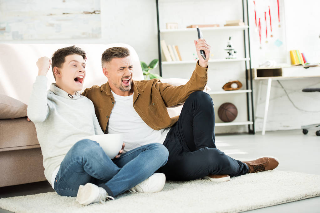 pai animado e filho adolescente assistindo jogo na tv e segurando tigela de pipocas em casa
 - Foto, Imagem
