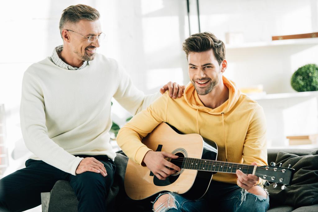 cheerful son playing acoustic guitar for mature father on weekend at home - Photo, Image