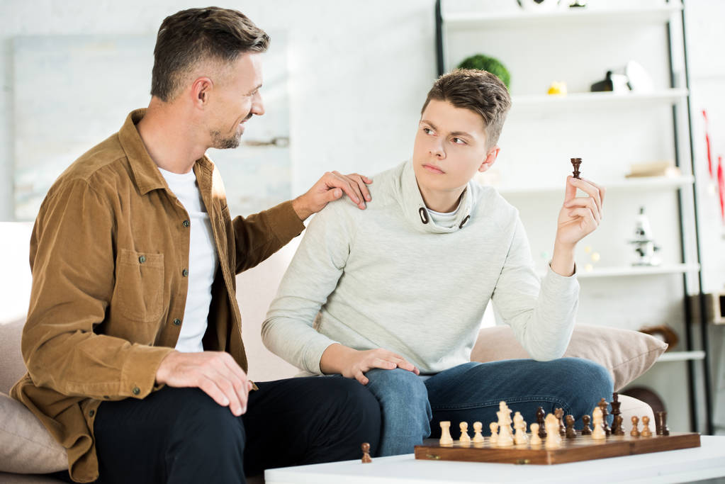 smiling father hugging teen son while he holding chess figure at home - Photo, Image