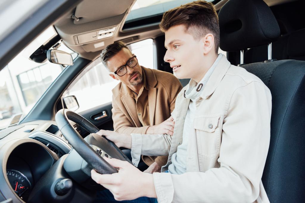 komea isä tukee teini-ikäistä poikaa opettaessaan häntä ajamaan autoa
 - Valokuva, kuva