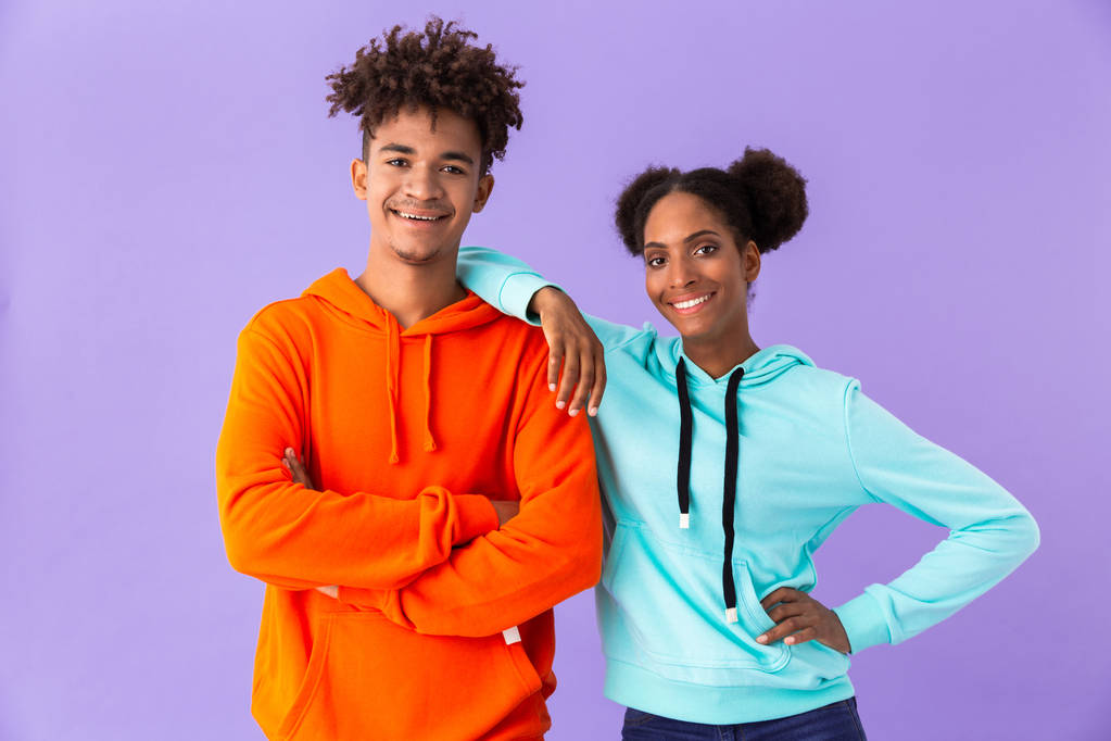 Foto van tiener Afrikaanse Amerikaanse echtpaar in kleurrijke kleding glimlachen terwijl je samen geïsoleerd over violette achtergrond - Foto, afbeelding