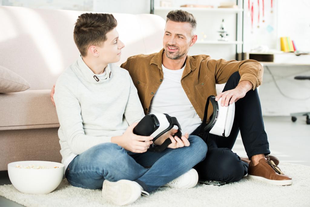 Lächelnder Vater und Teenager-Sohn, die Virtual-Reality-Headsets zu Hause halten und einander anschauen - Foto, Bild