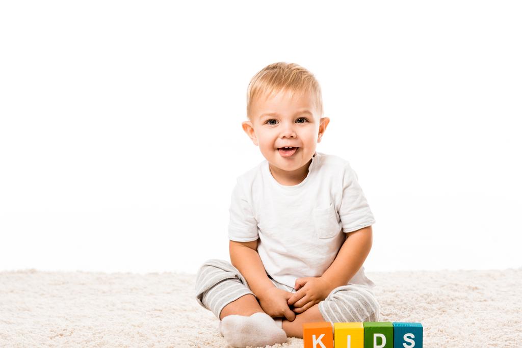 śmiejąc się toddler chłopiec siedzi na dywanie w pobliżu kolorowe kostki z literami na białym tle - Zdjęcie, obraz