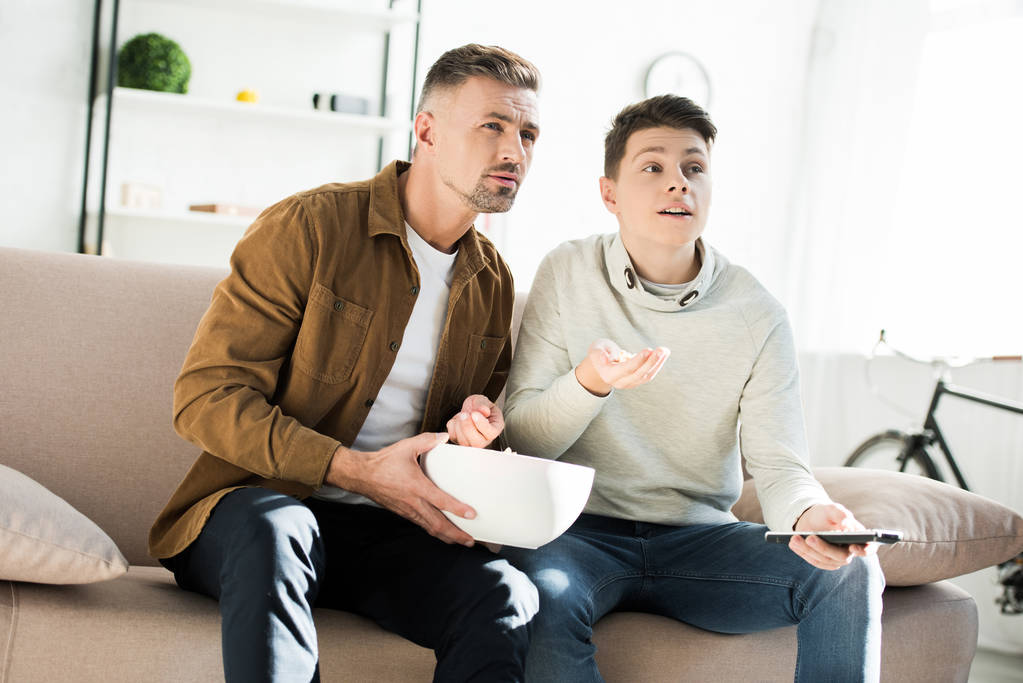 padre serio e hijo adolescente viendo la televisión y sosteniendo tazón de palomitas de maíz en casa
 - Foto, Imagen