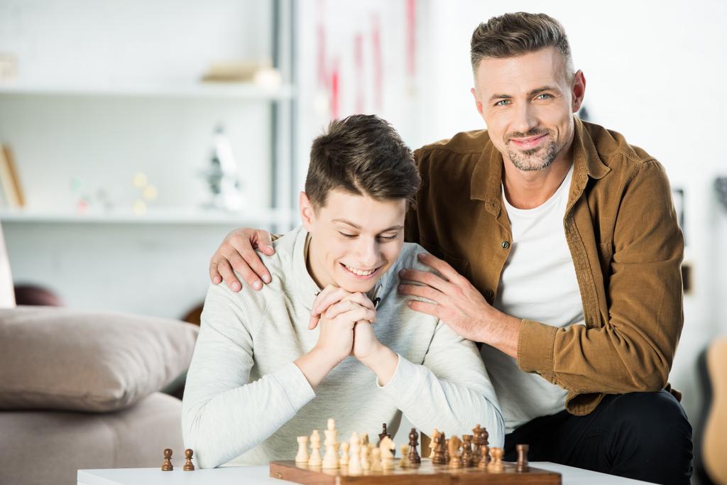 χαρούμενος ο πατέρας αγκαλιάζει teen γιος παίζοντας σκάκι στο σπίτι - Φωτογραφία, εικόνα