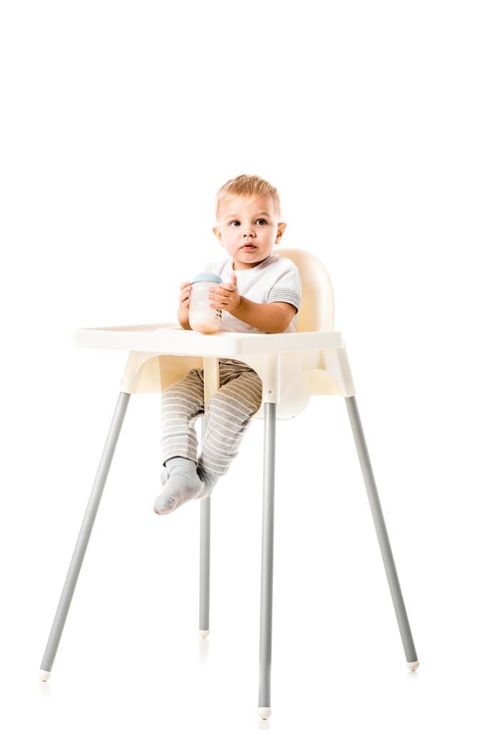 adorable toddler boy holding baby bottle and sitting in highchair isolated on white - Photo, Image