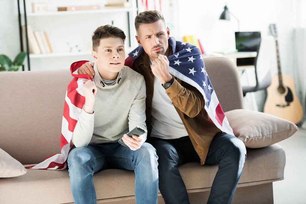 serious father and teen son wrapped in united states flag sitting on sofa and watching sport match - Photo, Image