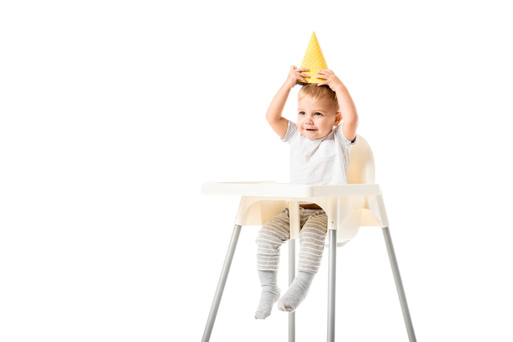 lindo niño sentado en trona y poniendo sombrero de fiesta amarillo en la cabeza aislado en blanco
 - Foto, Imagen