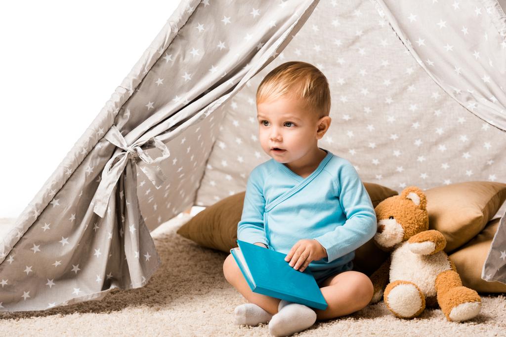 toddler boy holding blue book and sitting in wigwam with pillows and teddy bear isolated on white - Photo, Image