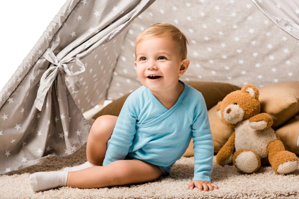 μικρό παιδί αγόρι κάθεται σε μωρό wigwam με μαξιλάρι και αρκουδάκι και γελώντας απομονωθεί σε λευκό - Φωτογραφία, εικόνα
