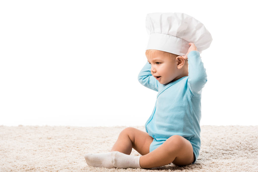 peuter jongen in koks hoed met handen op het hoofd zitten op tapijt geïsoleerd op wit - Foto, afbeelding