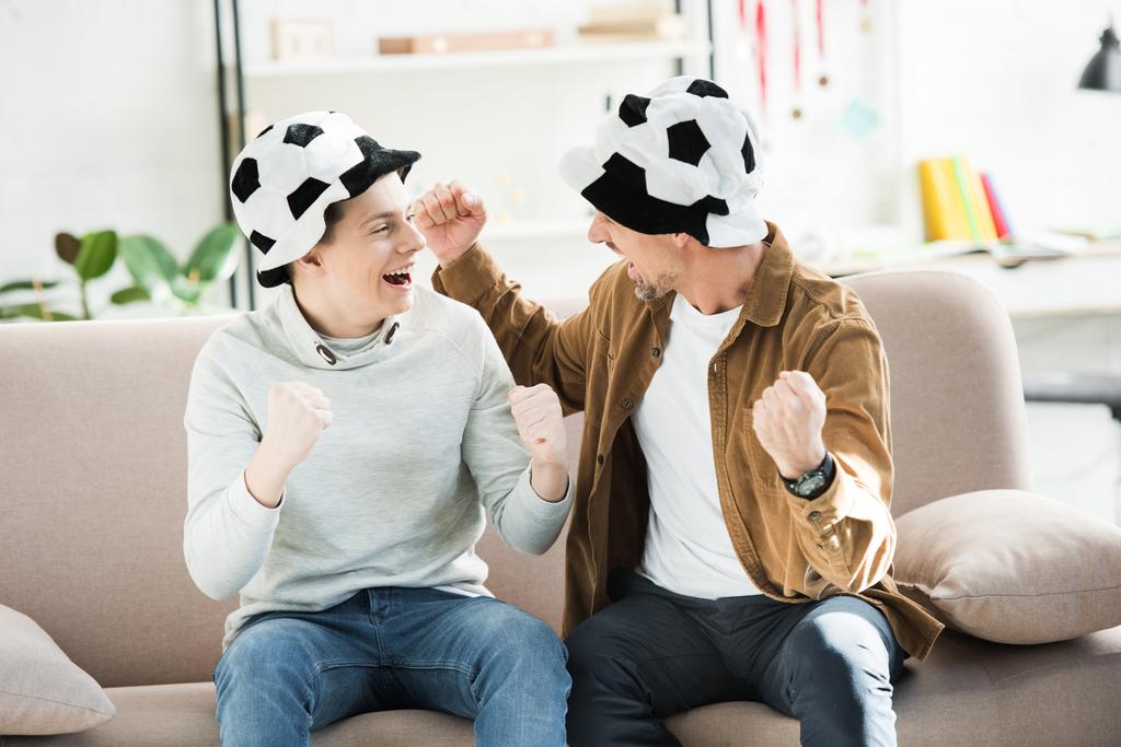aufgeregter Vater und Teenie-Sohn mit Fußballmützen, die sich zu Hause auf dem Sofa das Spiel anschauen und schreien und einander anschauen - Foto, Bild