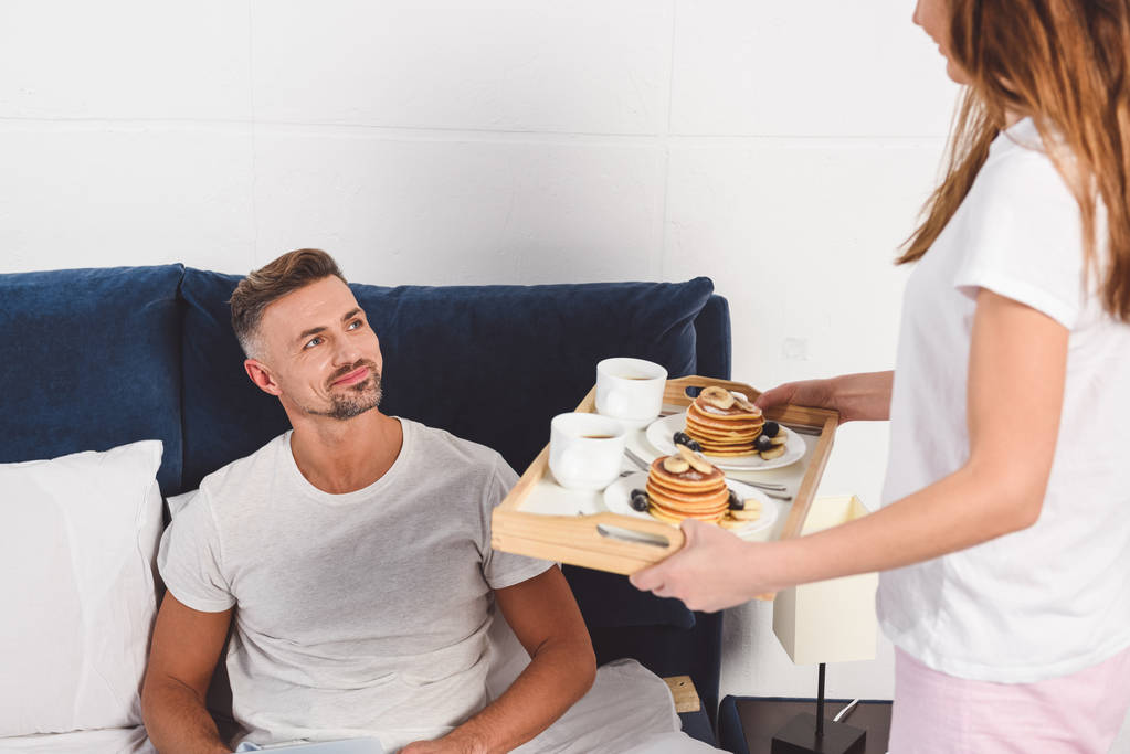 Esposa sosteniendo bandeja con desayuno y marido acostado en la cama
  - Foto, Imagen