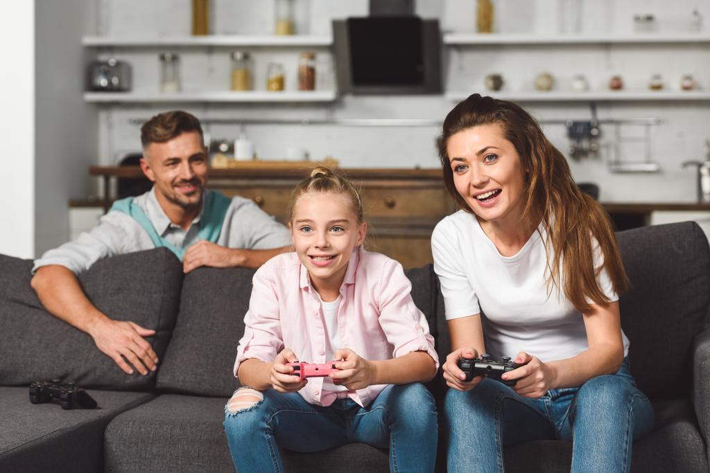 Lächelnde Mutter und Tochter beim Videospiel, während sich der Vater hinter dem Sofa versteckt - Foto, Bild