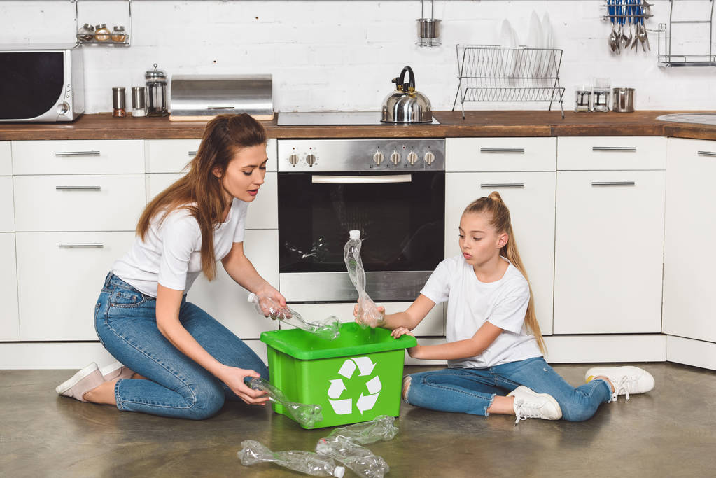 Mutter und Tochter sitzen in der Küche auf dem Boden und legen leere Plastikflaschen in Kartons mit Recyclingschild - Foto, Bild