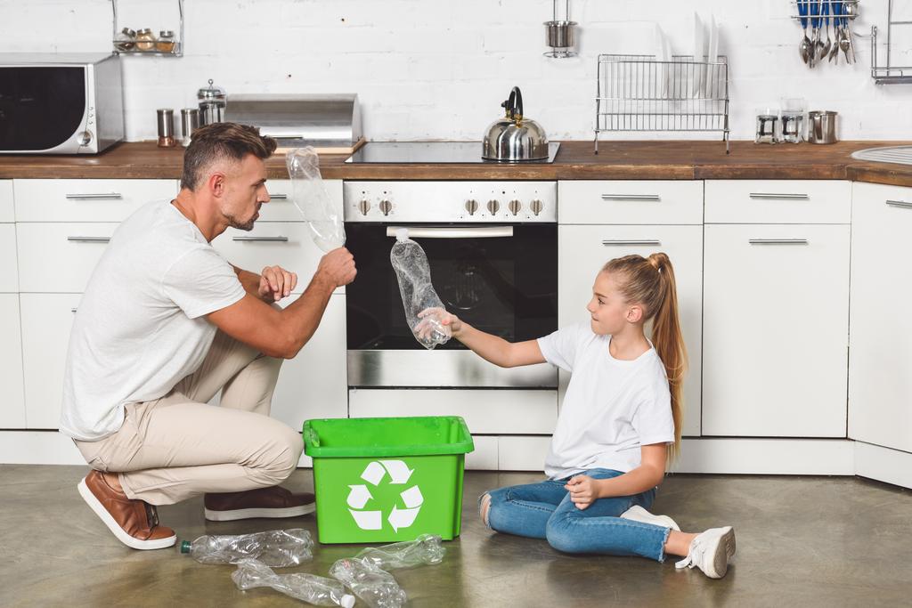 isä ja tytär istuvat lattialla keittiössä ja laittavat tyhjiä muovipulloja laatikkoon, jossa on kierrätyskyltti
 - Valokuva, kuva
