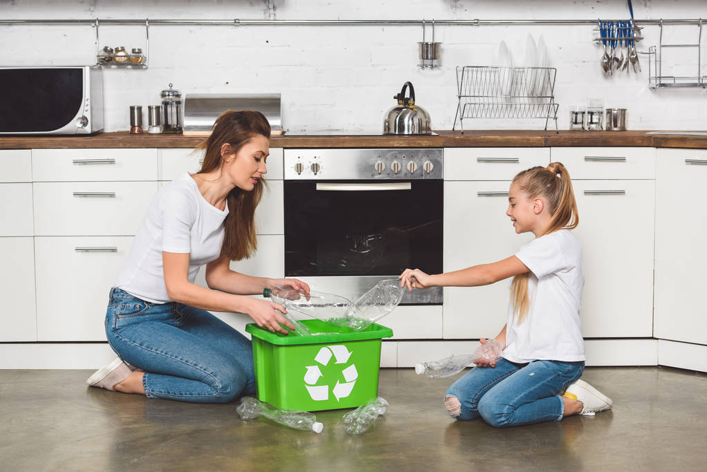 mãe e filha colocando garrafas de plástico vazias na caixa de reciclagem verde na cozinha
 - Foto, Imagem