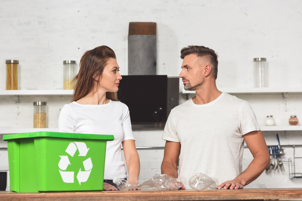 adult couple looking at each other with green recycle box and empty plastic bottles at kitchen table - Photo, Image