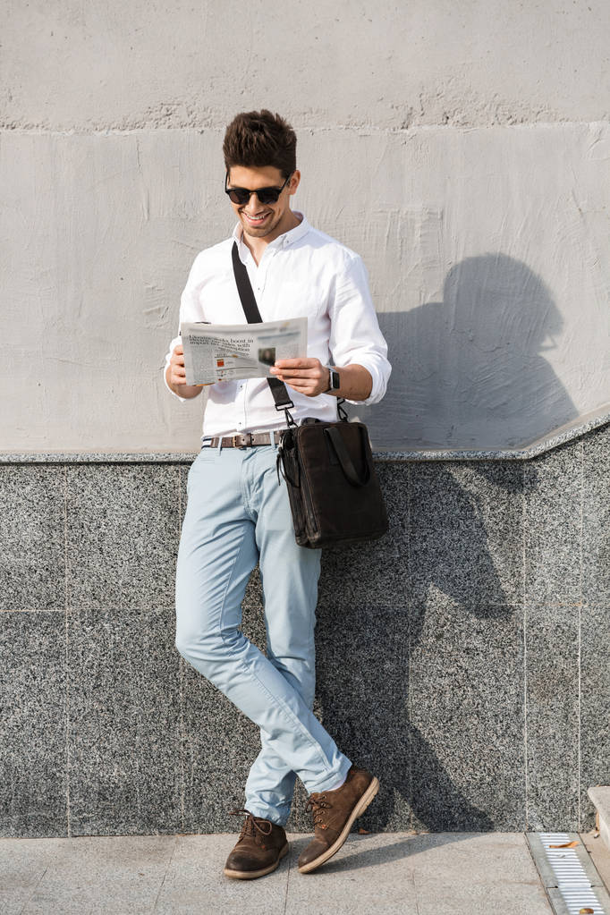 Fotografie mladého muže 30s čtenářský deník stoje podél venkovní zdi nosí sluneční brýle - Fotografie, Obrázek
