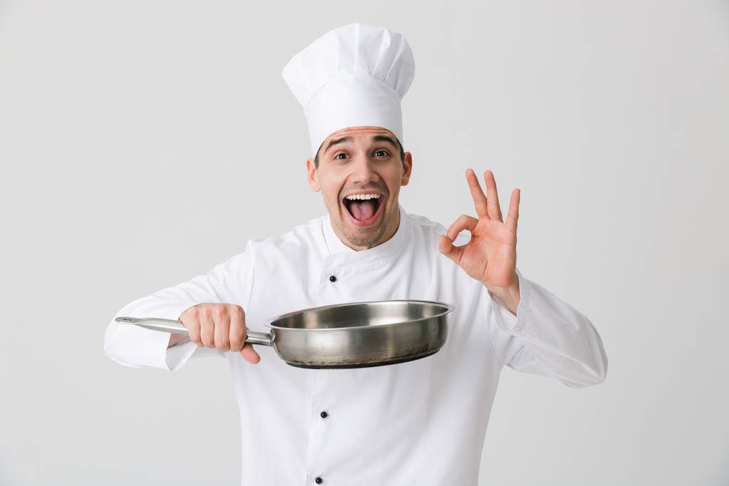 Изображение возбужденного эмоционального молодого человека шеф-повара в помещении на белом фоне, держащего сковородку
. - Фото, изображение